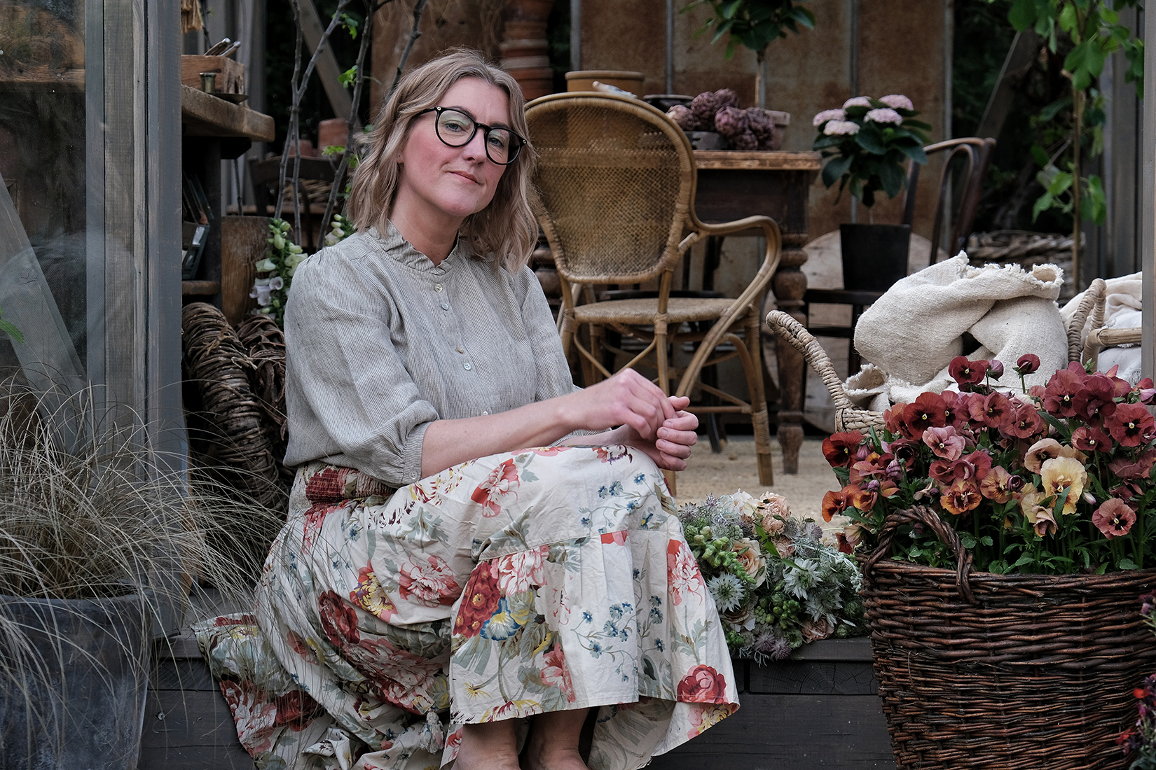 Linda Hansson i blommig kjol och linskjorta. Hon sitter bland sina vackra blomsterkreationer. 